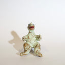 Ceramic sculpture of a sitting Godzilla, with its arms extended out as if for a hug. It has a variety of glazes, leaving it mostly a light mint green with brown undertones and speckles.