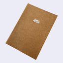 A brown kraft paper notebook with a small silver robot face illustration on the cover, shown at an angle against a light gray background.