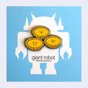 Three yellow and gold enamel pins shaped like tarts with smiling faces, arranged on a white card featuring a giant robot silhouette.