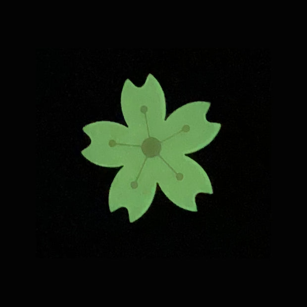 Die cut enamel pin of a stylized cherry blossom, with slightly pointed geometric petals and a white line art center. Pin is glowing in the dark.