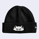 A black ribbed knit beanie hat featuring a white embroidered cartoon face with pointed ears and a small white script logo on the cuff, set against a light gray background.