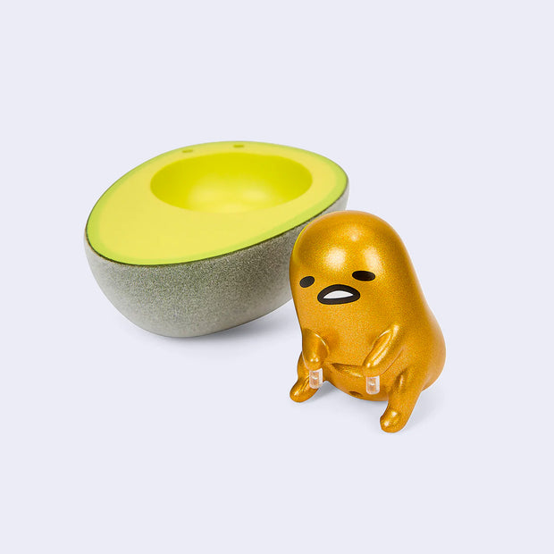 Vinyl figure of a metallic gold Gudetama, sitting on the floor next to half of a ripe avocado.