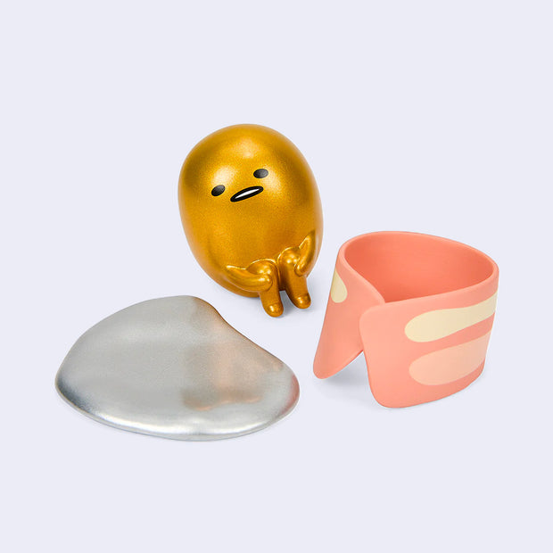 Vinyl figure of metallic gold Gudetama, sitting on the floor with its knees pulled into its stomach. Next to it is a silver metallic base and a piece of bacon, curled into itself.