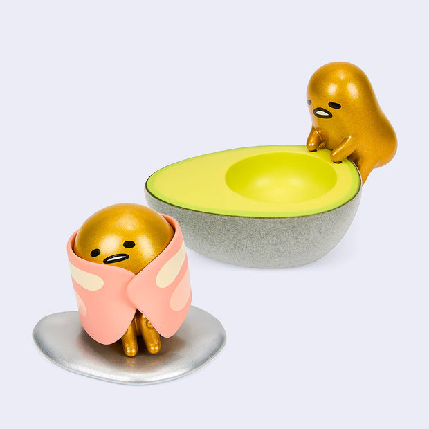 2 vinyl figures of Gudetama, a metallic gold egg yolk character with a comical expression. One figure climbs up into half of an avocado. The other figure sits with a piece of bacon wrapped around it, like a shawl.