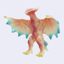 A small fantasy dragon figurine with spread wings, shown against a plain light gray background. The body is primarily pale pink and peach with blue-green wing edges and small yellow accents on the claws and tail tip.