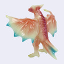 A small kaiju figurine with translucent wings and legs, featuring a gradient of peach, pink, purple, and turquoise blue, set against a plain light gray background.