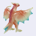 A small translucent kaiju figurine with wings spread, featuring a pink and peach body with blue-tipped wings and tail, shown against a plain light gray background.