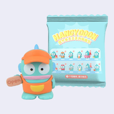 Small figurine of Hangyodon, dressed as a store worker with a sign that reads "open". It stands in front of its blind bag packaging.