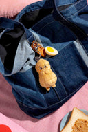 Keychain shaped like a cartoon peanut character attached to a denim bag on a pink surface with toast and peanut butter.