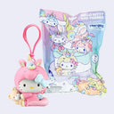 A Hello Kitty and Friends blind bag keychain charm shown next to its packaging. The charm features a pink unicorn-themed hood with a pink loop strap and a small teddy bear, while the packaging displays colorful illustrations of various Hello Kitty characters.