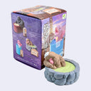 Small figurine of a monkey with brown fur and a pink face, resting in a spa with green water and stone walls. The monkey has a white folded towel atop its head and is positioned in front of its product packaging.