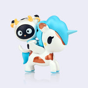Vinyl figure of Sanrio's Chococat, dressed in a full body cow outfit and riding atop the back of a white unicorn with brown metallic spots and a blue mane and tail.