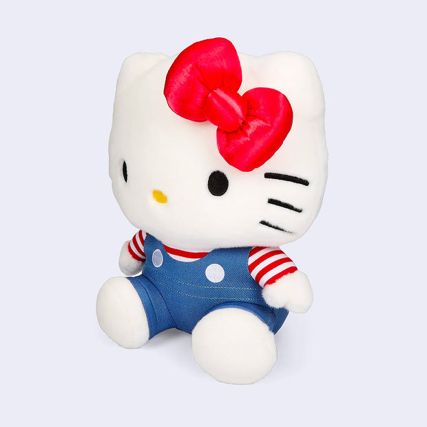 Plush doll of Hello Kitty, wearing blue overalls with a red and white striped shirt underneath. She has her iconic red bow. Her head is larger than her body.