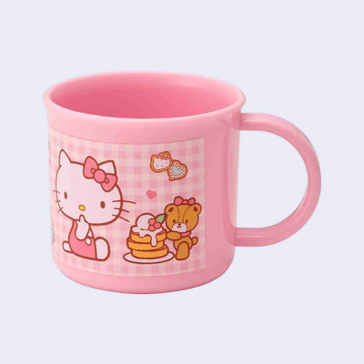 Small pink plastic cup with a mug handle featuring an illustration of Hello Kitty, sitting next to a stack of sweet pancakes that a stuffed bear holds. Background is pink gingham.