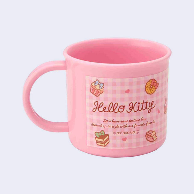 Small pink plastic cup with a mug handle featuring a written blurb about Hello Kitty, describing the character's personality. Background is pink gingham with decorative drawings of sweets.