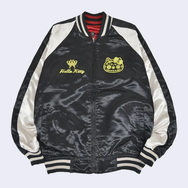 Black satin jacket with white side detailing and a gold graphic of Hello Kitty's head in a Day of the Dead skull pattern design. On the other side of the lapel is a gold butterfly and underneath cursive "Hello Kitty"