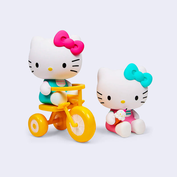 2 vinyl figures of Hello Kitty, dressed in her usual outfit but with pink and teal coloring. One figure rides a tricycle and the other sits on the ground and eats a vanilla ice cream cone.