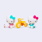 2 vinyl figures of Hello Kitty, dressed in her usual outfit but with pink and teal coloring. One figure rides a tricycle and the other sits on the ground and eats a vanilla ice cream cone.