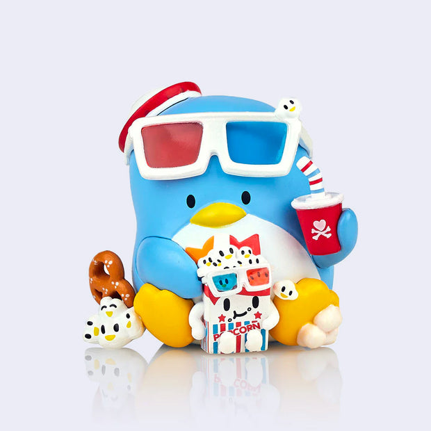 Vinyl figure of Sanrio's Tuxedosam, sitting on the ground with 3D glasses atop his head. He holds a soda in one hand, and a box of popcorn between his legs with some kernels near his feet.