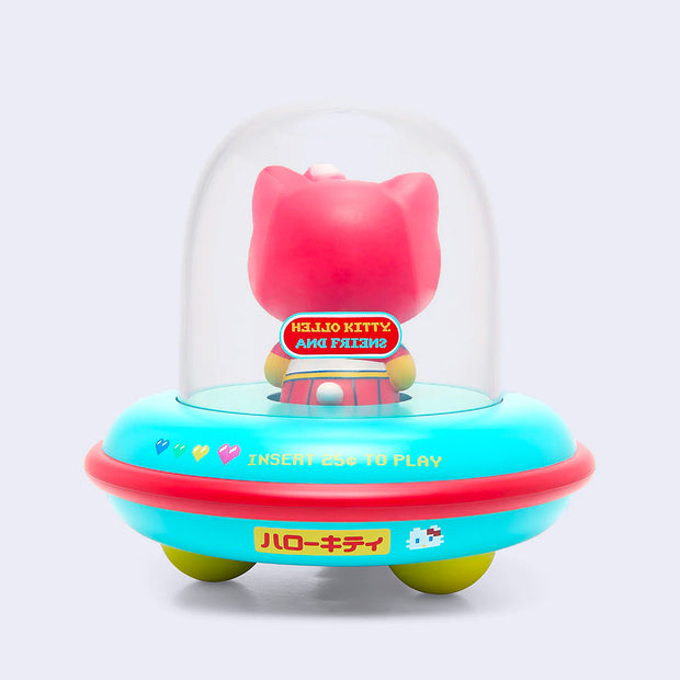 Back view of vinyl figure of Hello Kitty, dressed in a red space suit. She sits inside of a UFO with a donut shaped body that is teal and reads "Insert 25 cents to Play." On the UFO body is an 8 bit style drawing of Hello Kitty.