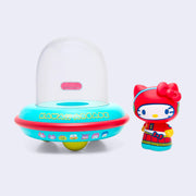 Vinyl figure of Hello Kitty, dressed in a red space suit. She stands outside of a UFO with a donut shaped body that is teal and reads "Kawaii Arcade." On the UFO body are 8 bit style drawings of Sanrio Characters.