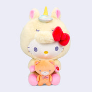 Plush of Hello Kitty, dressed in a full body fluffy light yellow unicorn costume with a horn and iridescent bow. She holds in her hands a brown teddy bear.