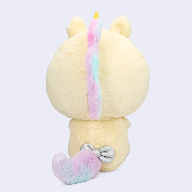 Plush of Hello Kitty, dressed in a full body fluffy light yellow unicorn costume with a horn and iridescent bow. She holds in her hands a brown teddy bear. Back angle.