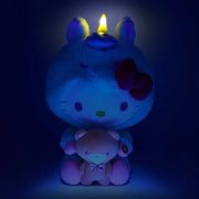 Plush of Hello Kitty, dressed in a full body fluffy light yellow unicorn costume with a horn and iridescent bow. She holds in her hands a brown teddy bear. Her horn emits a soft glow.