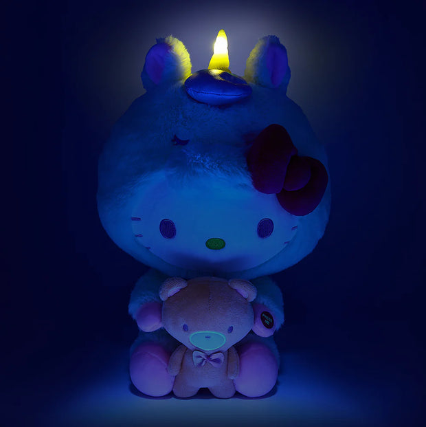 Plush of Hello Kitty, dressed in a full body fluffy light yellow unicorn costume with a horn and iridescent bow. She holds in her hands a brown teddy bear. Her horn emits a soft glow.