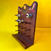 Wooden thread holder, with many pegs to hold many spools of thread, with a retro style pattern painted atop it of 2 pink birds and florals.