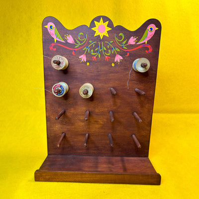 Wooden thread holder, with many pegs to hold many spools of thread, with a retro style pattern painted atop it of 2 pink birds and florals.