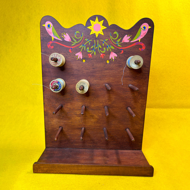 Wooden thread holder, with many pegs to hold many spools of thread, with a retro style pattern painted atop it of 2 pink birds and florals.
