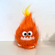 Very fluffy orange plush in the shape of a flame, with 2 large plastic eyes, an underbite and small feet buried beneath its fur. 