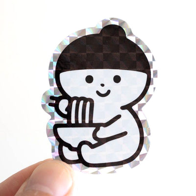 Holographic sticker of a small character sitting and eating a bowl of noodles with chopsticks.