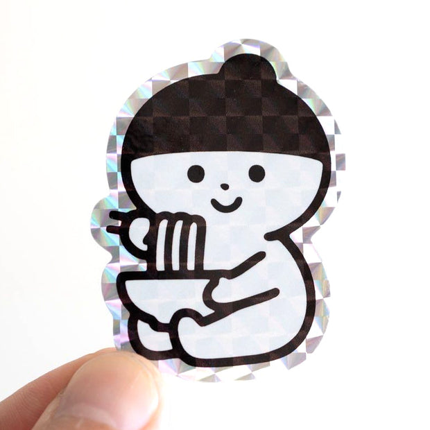 Holographic sticker of a small character sitting and eating a bowl of noodles with chopsticks.