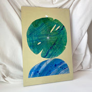 Collage style painting on solid taupe background of a large, flat green lily pad stacked atop of a blue rock. Lily pad and rock both have bold abstract marbling patterns and a small white butterfly rests atop the lily pad. Displayed at an angle.