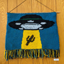 Wall hanging crochet tapestry of a large UFO abducting a cactus.
