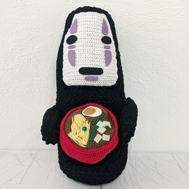 Crocheted sculpture of No Face from Spirited Away holding a red bowl of ramen, with noodles, egg, tofu and green onion.