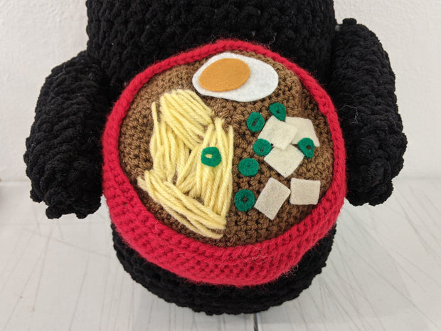 Close up of crocheted sculpture of No Face from Spirited Away holding a red bowl of ramen, with noodles, egg, tofu and green onion.