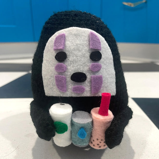 Crochet plush of No Face, from Spirited Away holding 3 drinks: a coffee, a water and a strawberry boba.