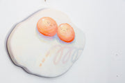 Die cut canvas of a pair of 2 eggs sharing a single egg white. The yolks are touching and have small faces. The egg white contains a subtle polka dot and scribble design.