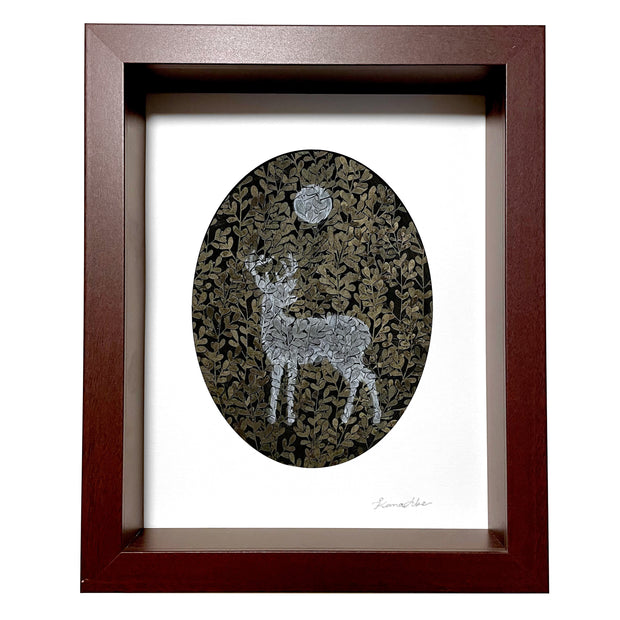 Paper cutting of many yellowish green leaves, with a silhouette of a deer painted onto the trees in a light blue. Above, using the same technique is a moon. Piece is in thick wooden frame.