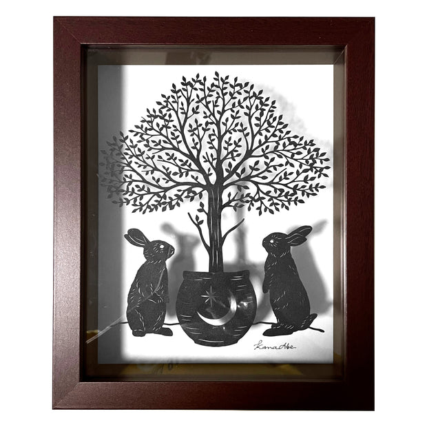 Paper cutting art from black paper, of 2 rabbits standing on their hind legs around a potted tree, with many individual leaves. Piece is in a thick wooden shadowbox frame.