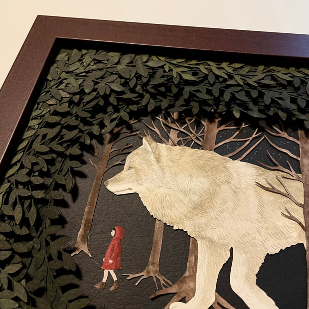 Assembled paper cutting diorama of a large white wolf, following a small girl in a red coat through a forest. They are framed by many cut leaves revealing the scene. Close up showing 3 dimensionality.