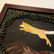 Assembled paper cutting diorama of a fox, jumping over a mountain range and against a starry night sky. It is framed by many cut leaves, revealing the scene. Close up to show 3 dimensionality.