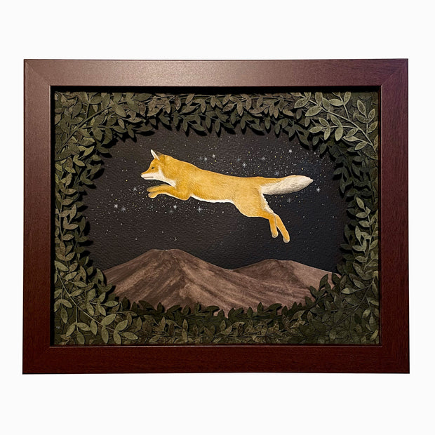 Assembled paper cutting diorama of a fox, jumping over a mountain range and against a starry night sky. It is framed by many cut leaves, revealing the scene. Piece is in open shadowbox style wooden frame.