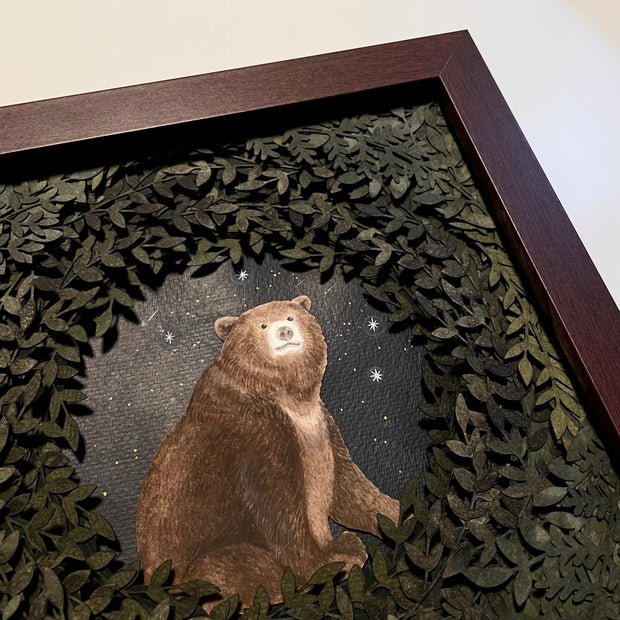 Layered cut paper diorama style sculpture in an open wooden frame, with many layers of leaves revealing a small scene of a bear sitting and smiling slightly. Shooting stars fall behind it. Close up to show 3 dimensionality.