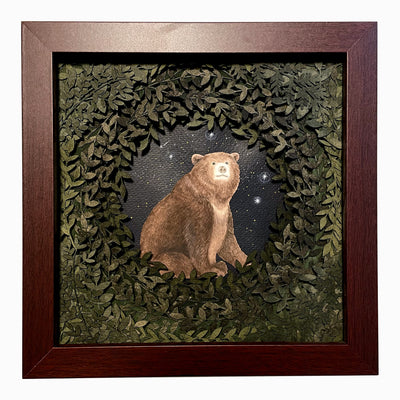 Layered cut paper diorama style sculpture in an open wooden frame, with many layers of leaves revealing a small scene of a bear sitting and smiling slightly. Shooting stars fall behind it.