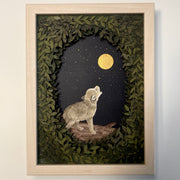 Framed paper cutting artwork of a small wolf, standing and howling up at the moon, gold in the dark night sky. Piece is framed by an oval assembling of cut leaves.