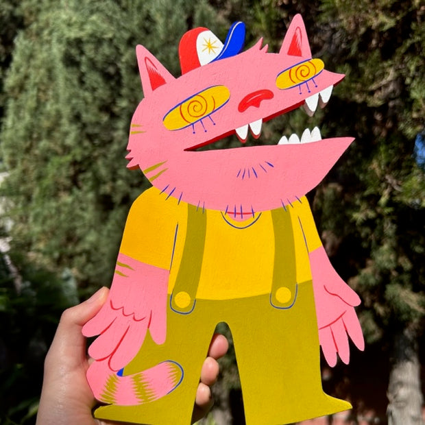 Die cut wooden sculpture of a pink cat, with a dazed expression and an open mouth. It has large human hands and wears olive green overalls with a yellow shirt under.